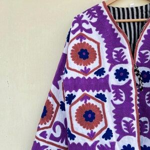 May include: A white jacket with purple and blue embroidery. The jacket has a geometric pattern with floral motifs. The jacket is lined with black and white striped fabric.
