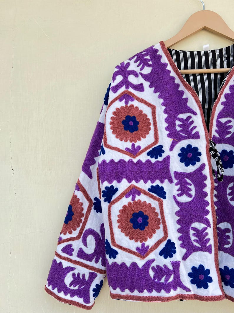 May include: A white jacket with purple and blue embroidery. The jacket has a geometric pattern with floral motifs. The jacket is lined with black and white striped fabric.