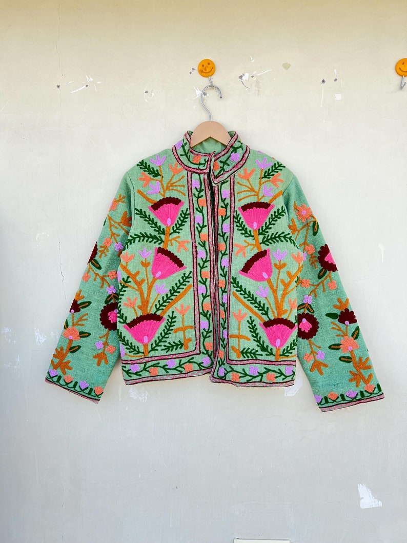 May include: A green embroidered jacket with floral patterns in pink, orange, and purple. The jacket has a stand-up collar and a button closure.