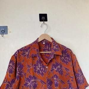 May include: Orange button-down shirt with a purple floral pattern. The short-sleeved shirt is hanging on a wooden hanger. The shirt has a classic collar and a single chest pocket. The floral design is a repeating pattern of large flowers and leaves.
