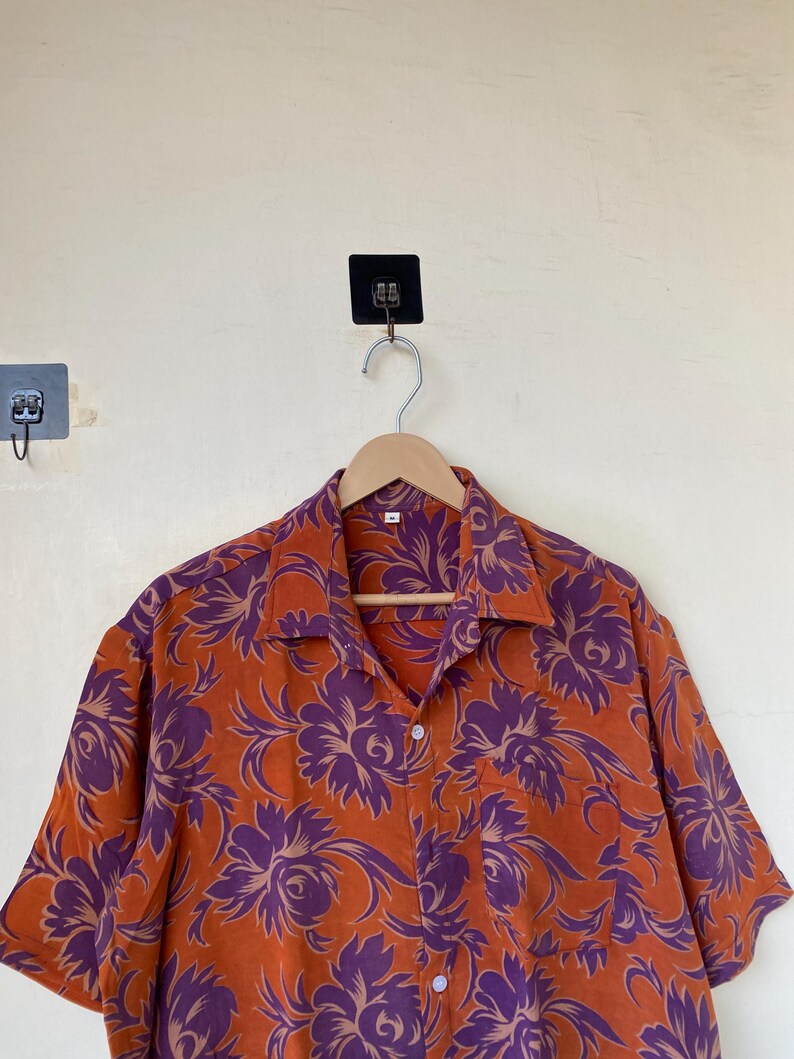 May include: Orange button-down shirt with a purple floral pattern. The short-sleeved shirt is hanging on a wooden hanger. The shirt has a classic collar and a single chest pocket. The floral design is a repeating pattern of large flowers and leaves.