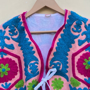 Puede incluir: Un chaleco rosa bordado con motivos florales azules, verdes y rosas. El chaleco tiene un cierre de corbata blanca.