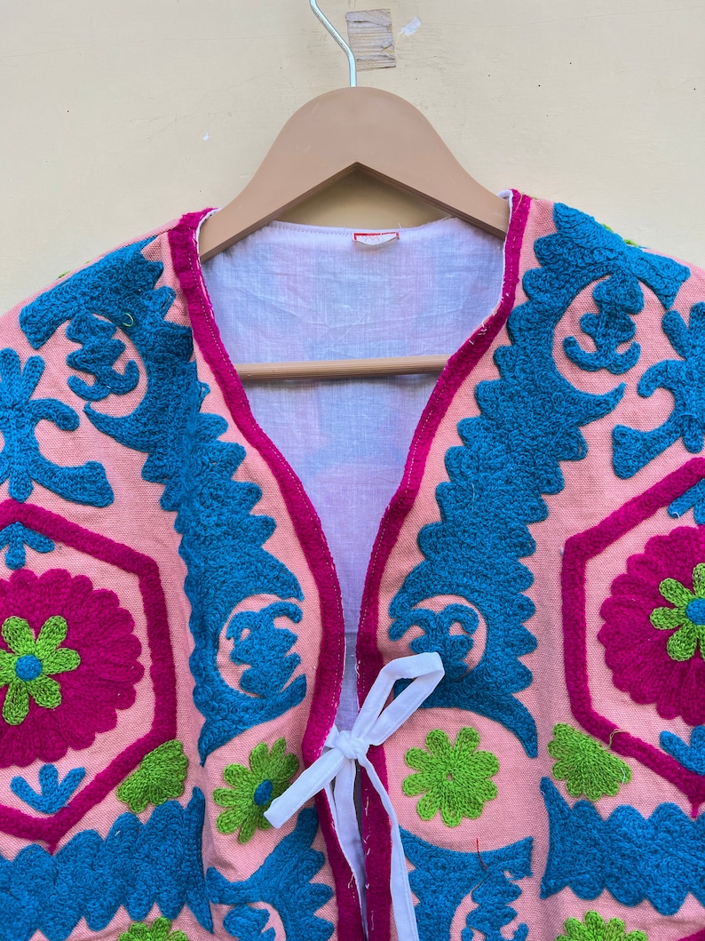 Puede incluir: Un chaleco rosa bordado con motivos florales azules, verdes y rosas. El chaleco tiene un cierre de corbata blanca.
