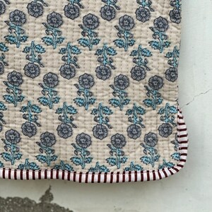 May include: A quilted textile with a floral pattern in shades of gray and blue on a cream background. The edge is trimmed with a striped border in maroon and white. The stitching is visible, adding texture to the fabric.