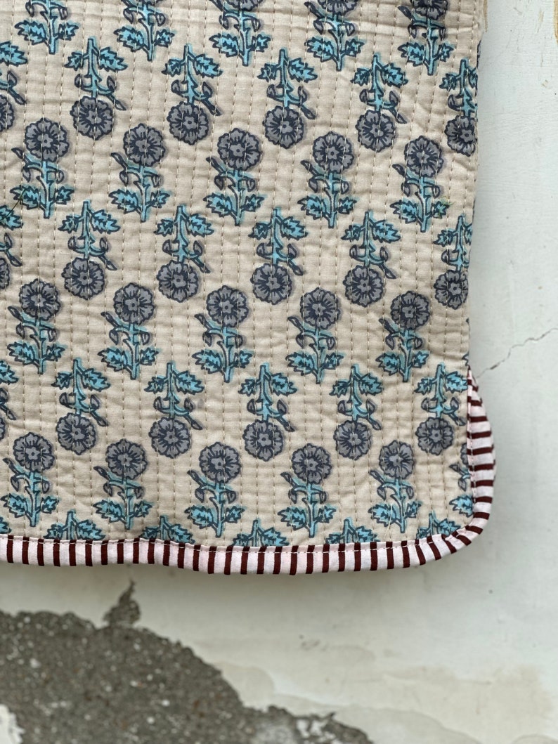 May include: A quilted textile with a floral pattern in shades of gray and blue on a cream background. The edge is trimmed with a striped border in maroon and white. The stitching is visible, adding texture to the fabric.