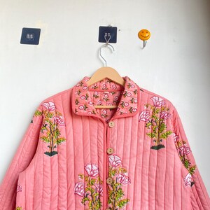 Pu&ograve; includere: Giacca trapuntata color corallo con stampa floreale. La giacca ha una chiusura con bottoni, due tasche anteriori e un colletto. Il design floreale comprende fiori rosa e foglie verdi. La giacca &egrave; appesa a un'appendiabiti di legno.