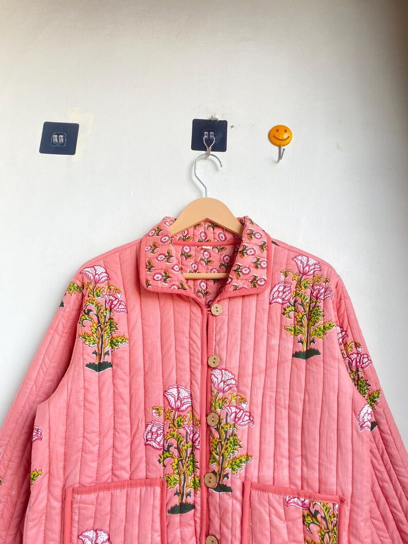 Pu&ograve; includere: Giacca trapuntata color corallo con stampa floreale. La giacca ha una chiusura con bottoni, due tasche anteriori e un colletto. Il design floreale comprende fiori rosa e foglie verdi. La giacca &egrave; appesa a un'appendiabiti di legno.