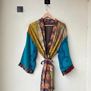 May include: A kimono-style robe featuring a patchwork design with vibrant panels of yellow, teal, and floral patterns. The garment has wide sleeves, a wrap-front closure with a matching fabric tie belt, and a contrasting black and white striped lining.