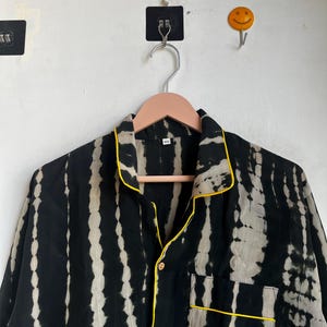Może przedstawiać: Czarno-biała koszula piżamowa w stylu tie-dye z ż&oacute;łtymi wykończeniami. Koszula ma zapięcie na guziki z przodu, kieszeń na piersi i długie rękawy. Koszula wisi na r&oacute;żowym drewnianym wieszaku.