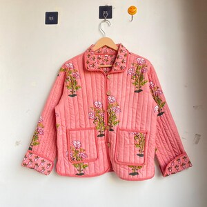 Pu&ograve; includere: Giacca trapuntata color corallo con ricami floreali. La giacca presenta un collo, due tasche anteriori e bottoni in legno. Il design floreale comprende fiori rosa e foglie verdi.