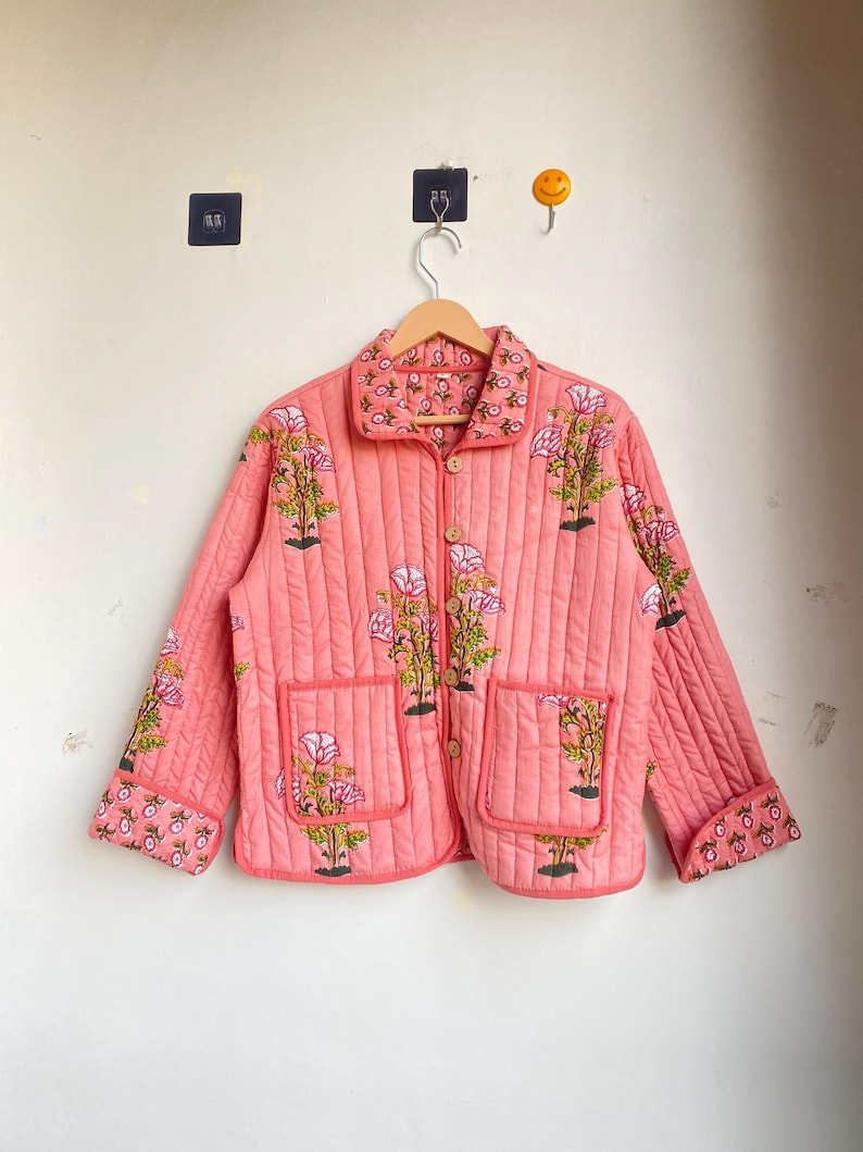 Pu&ograve; includere: Giacca trapuntata color corallo con ricami floreali. La giacca presenta un collo, due tasche anteriori e bottoni in legno. Il design floreale comprende fiori rosa e foglie verdi.