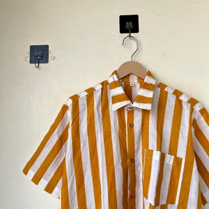 May include: A short-sleeved shirt with vertical stripes in mustard yellow and white. The shirt has a button-down front, a chest pocket, and a collar. It is hanging on a wooden hanger.