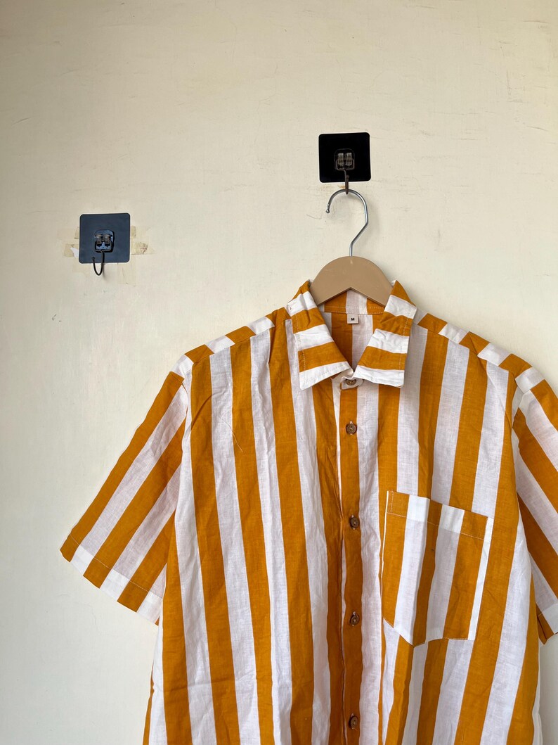 May include: A short-sleeved shirt with vertical stripes in mustard yellow and white. The shirt has a button-down front, a chest pocket, and a collar. It is hanging on a wooden hanger.
