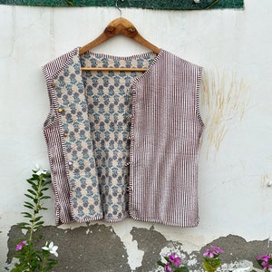 May include: A sleeveless vest with a striped exterior in white and dark brown, and a floral patterned interior in blue and beige. The vest is buttoned and hangs on a wooden hanger against a white wall.