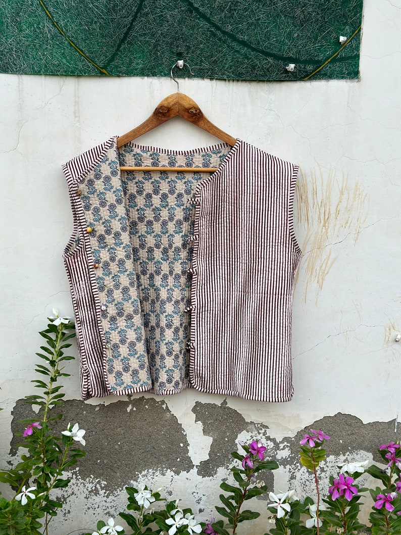 May include: A sleeveless vest with a striped exterior in white and dark brown, and a floral patterned interior in blue and beige. The vest is buttoned and hangs on a wooden hanger against a white wall.