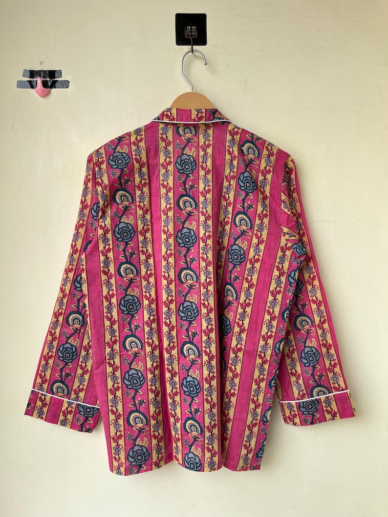 May include: A pink pajama top with vertical stripes and floral patterns in blue and beige. The long-sleeved top has white trim along the collar and cuffs, hanging on a wooden hanger.