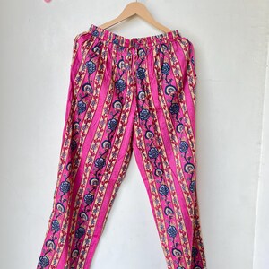 May include: Pink pajama pants with vertical stripes and floral patterns in blue, beige, and white. The pants are hanging on a wooden hanger against a white background. The pants have a drawstring waist and white trim.
