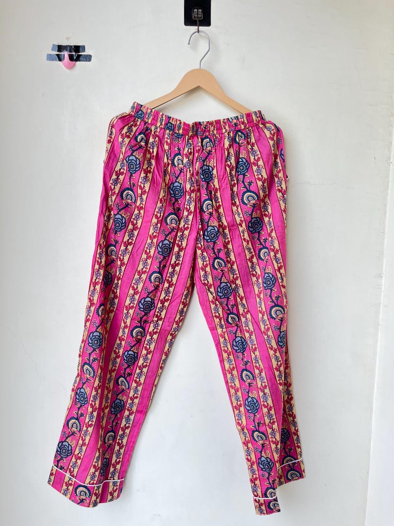 May include: Pink pajama pants with vertical stripes and floral patterns in blue, beige, and white. The pants are hanging on a wooden hanger against a white background. The pants have a drawstring waist and white trim.