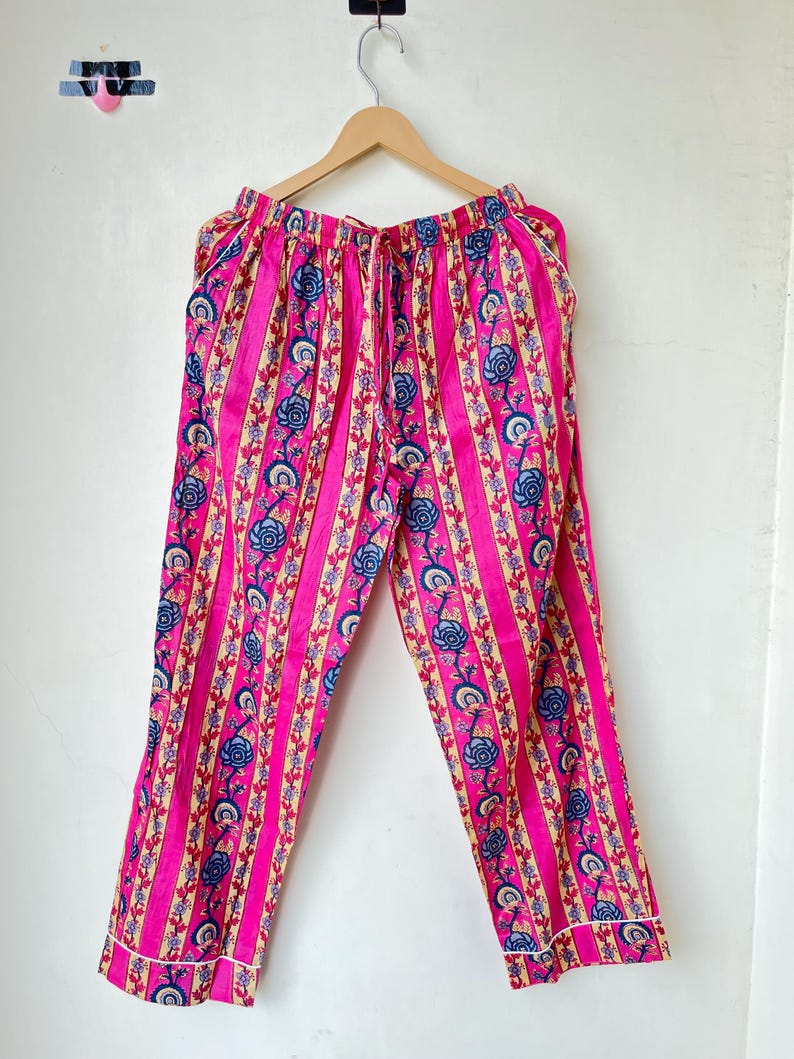 May include: Pink pajama pants with vertical stripes and floral patterns in blue, beige, and red. The pants have a drawstring waist and white trim along the cuffs. The fabric appears to be lightweight.