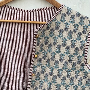 May include: A quilted vest with a floral pattern on one side and a striped pattern on the other. The floral side features blue flowers on a cream background. The striped side has vertical burgundy and white stripes. The vest is fastened with small, decorative buttons.
