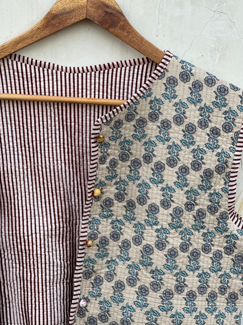May include: A quilted vest with a floral pattern on one side and a striped pattern on the other. The floral side features blue flowers on a cream background. The striped side has vertical burgundy and white stripes. The vest is fastened with small, decorative buttons.