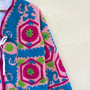 Puede incluir: Una chaqueta rosa con patrones florales bordados en azul, verde y rosa. La chaqueta tiene un cierre de corbata en el cuello.