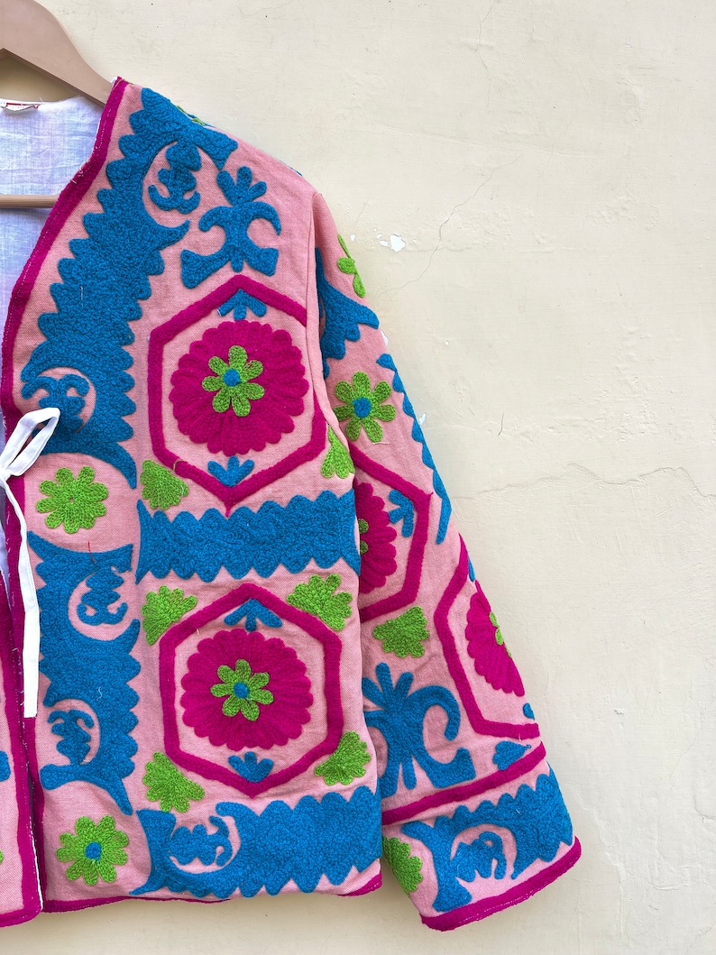 Puede incluir: Una chaqueta rosa con patrones florales bordados en azul, verde y rosa. La chaqueta tiene un cierre de corbata en el cuello.