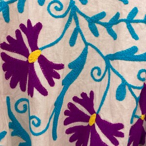 May include: White cotton fabric with colorful embroidery. The embroidery features a floral pattern with purple, blue, yellow, and orange thread.