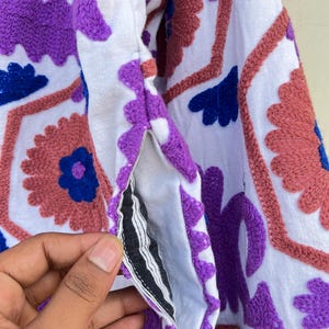 May include: A white fabric garment with purple, pink, and blue embroidered floral designs. The fabric has a textured, slightly fuzzy surface.
