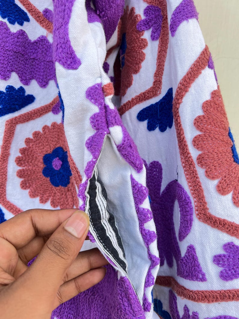 May include: A white fabric garment with purple, pink, and blue embroidered floral designs. The fabric has a textured, slightly fuzzy surface.