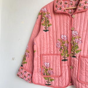 Pu&ograve; includere: Giacca rosa trapuntata con motivo floreale. La giacca presenta un colletto, due tasche frontali e bottoni in legno. Il motivo floreale include fiori rosa e foglie verdi.