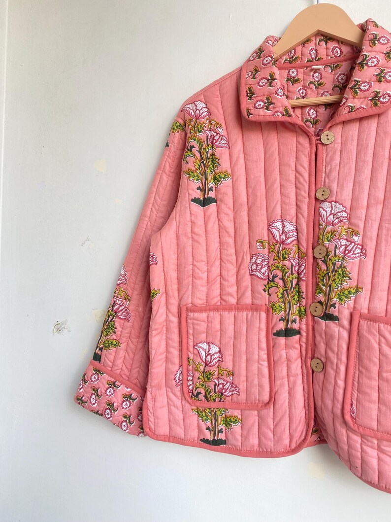 Pu&ograve; includere: Giacca rosa trapuntata con motivo floreale. La giacca presenta un colletto, due tasche frontali e bottoni in legno. Il motivo floreale include fiori rosa e foglie verdi.