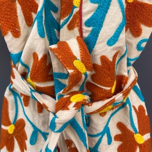 May include: A white cotton robe with a floral embroidery design. The robe features a tie belt and a collar. The embroidery is in shades of orange, blue, and purple.