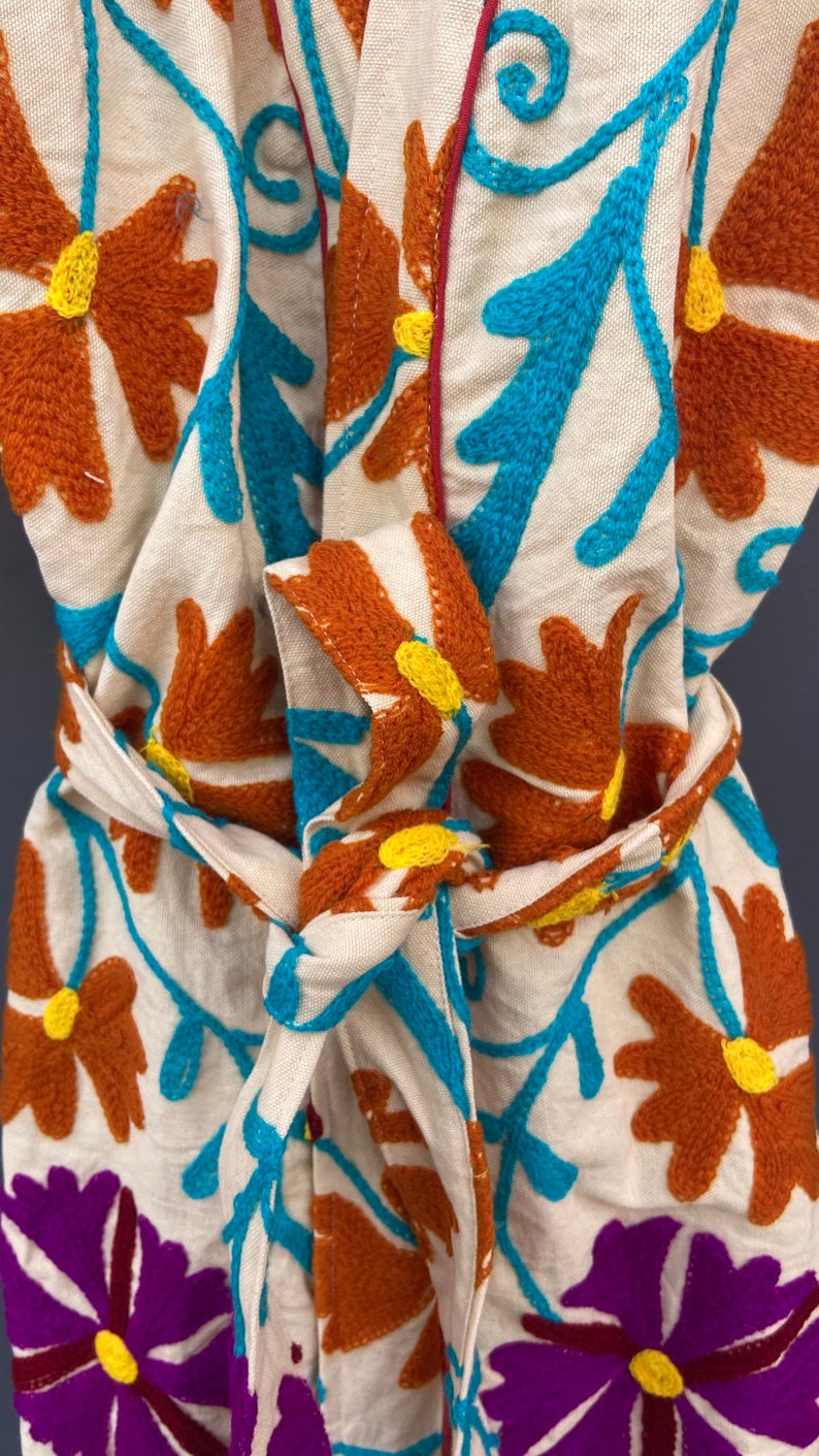 May include: A white cotton robe with a floral embroidery design. The robe features a tie belt and a collar. The embroidery is in shades of orange, blue, and purple.