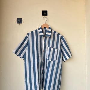 May include: Short-sleeved shirt with vertical blue and white stripes. The shirt has a button-down front, a chest pocket, and a classic collar. The shirt is hanging on a wooden hanger.