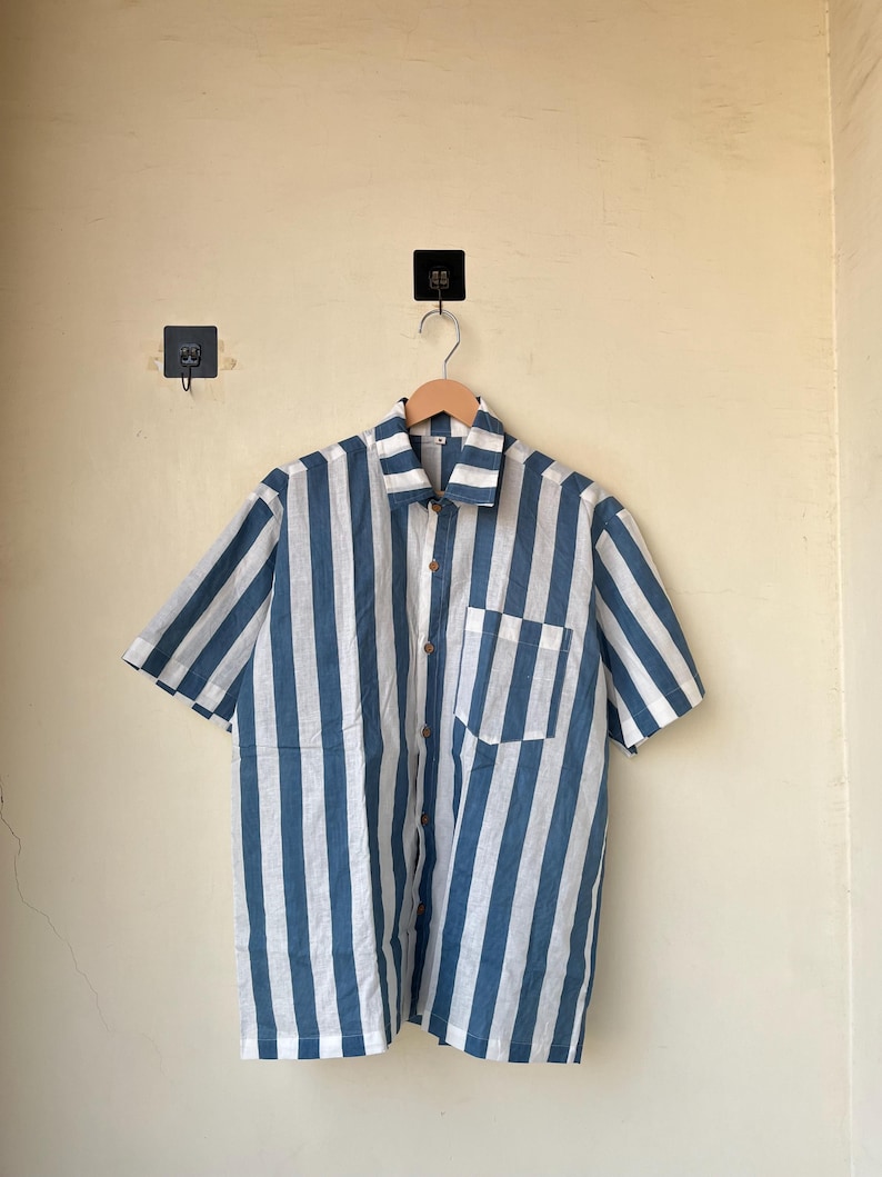 May include: Short-sleeved shirt with vertical blue and white stripes. The shirt has a button-down front, a chest pocket, and a classic collar. The shirt is hanging on a wooden hanger.