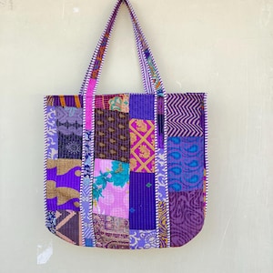 May include: A purple patchwork tote bag with a white and purple striped trim. The bag is made of different fabrics with various patterns and colours, including pink, blue, and gold.