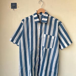 May include: Short-sleeved shirt with vertical blue and white stripes. The shirt has a button-down front, a chest pocket, and wooden buttons. The shirt is hanging on a wooden hanger.