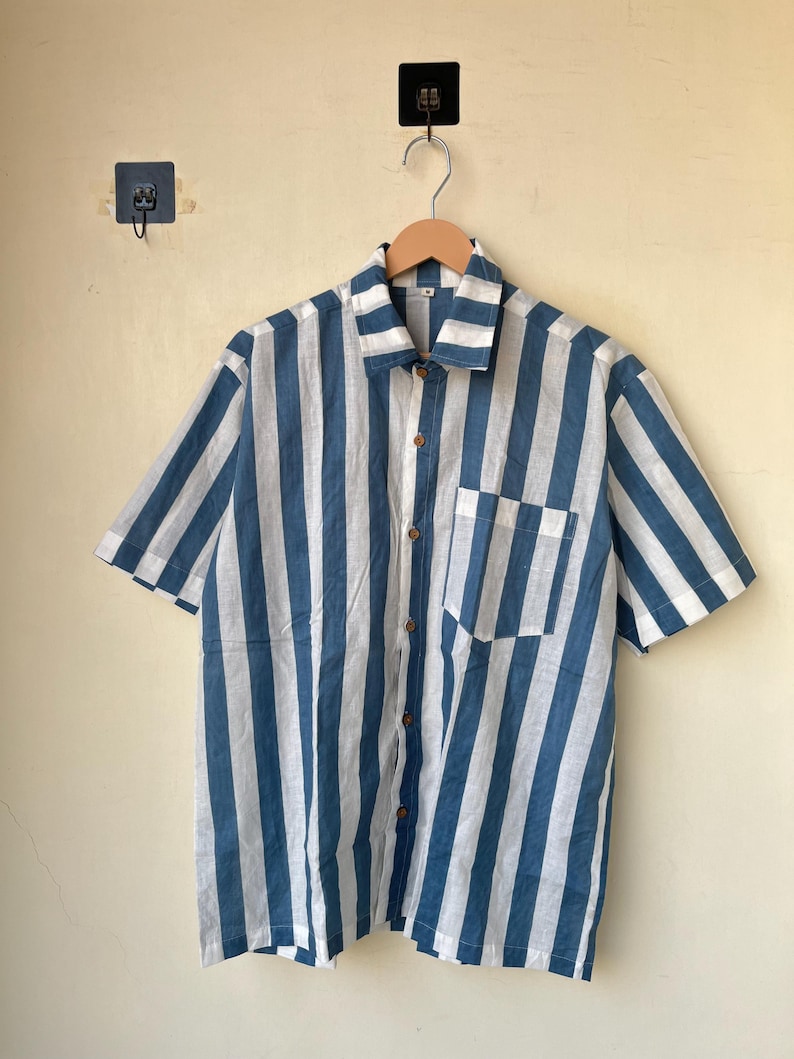 May include: Short-sleeved shirt with vertical blue and white stripes. The shirt has a button-down front, a chest pocket, and wooden buttons. The shirt is hanging on a wooden hanger.