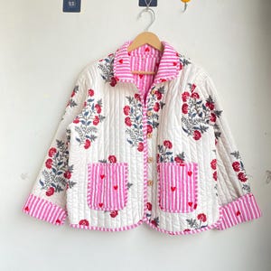 以下が含まれることがあります： 花柄とピンクのストライプのアクセントが施されたキルティングジャケット。赤い花、青い葉、ハートのディテールが施されたピンクのストライプのポケットが特徴です。スカラップの裾と木製のボタンが付いています。