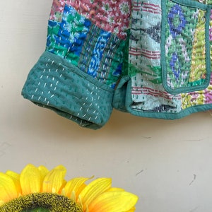 Puede incluir: Una chaqueta acolchada verde con un dise&ntilde;o de patchwork que presenta varios patrones florales y geom&eacute;tricos. La chaqueta tiene manga larga y cierre de bot&oacute;n.