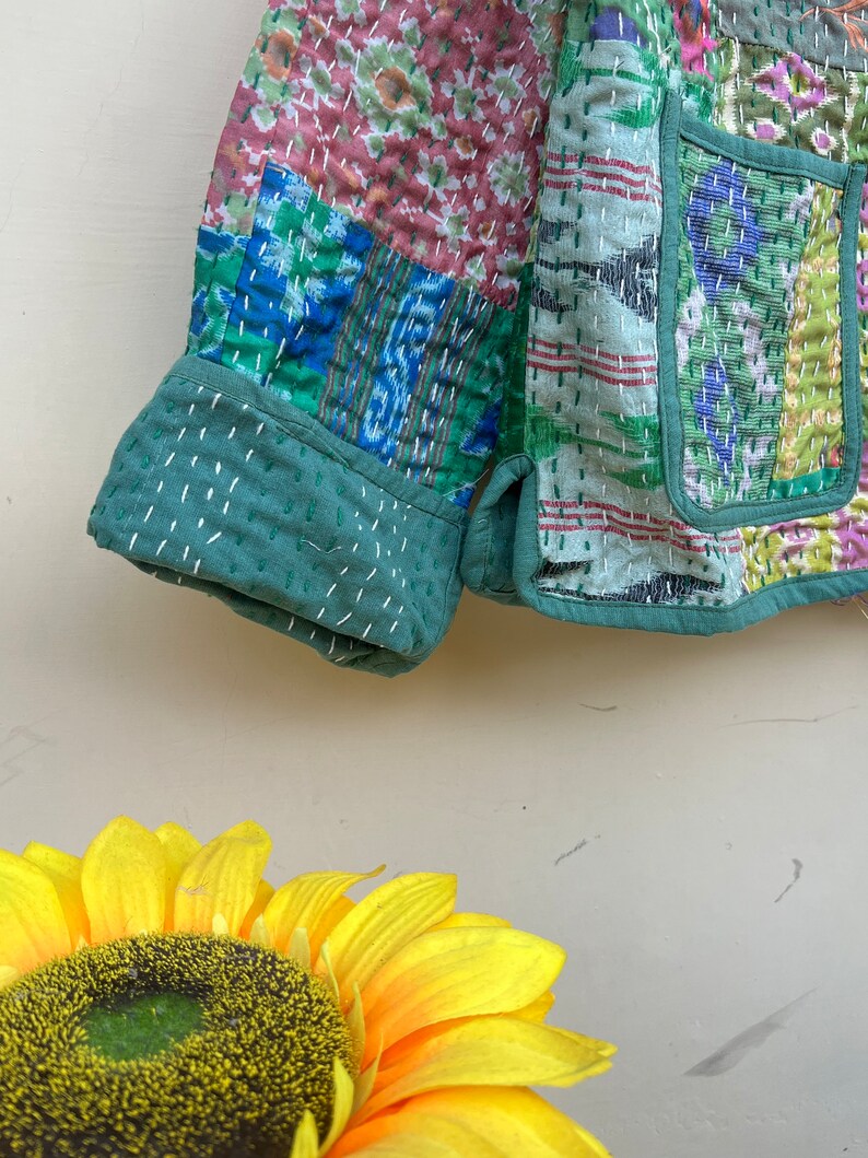 Puede incluir: Una chaqueta acolchada verde con un dise&ntilde;o de patchwork que presenta varios patrones florales y geom&eacute;tricos. La chaqueta tiene manga larga y cierre de bot&oacute;n.