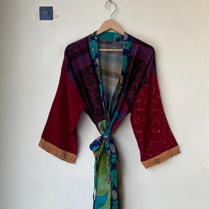 May include: A long-sleeved kimono-style robe featuring a patchwork design with deep red, blue, and green panels. The garment includes a tie-waist belt, wide sleeves with tan cuffs, and a multi-colored floral and paisley pattern throughout the fabric.