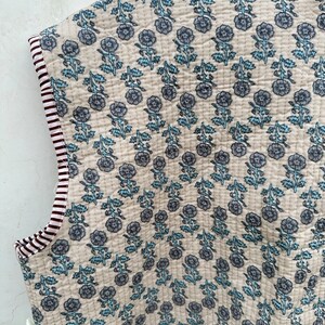 May include: Cream-colored quilted fabric with a floral pattern in shades of blue. The edges are trimmed with a striped border in burgundy and white. The fabric is likely used for a vest or similar garment.