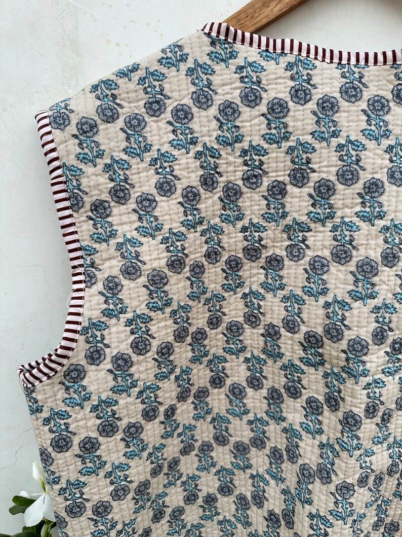 May include: Cream-colored quilted fabric with a floral pattern in shades of blue. The edges are trimmed with a striped border in burgundy and white. The fabric is likely used for a vest or similar garment.