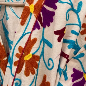 May include: A white cotton robe with a colorful floral embroidery design. The embroidery features turquoise, purple, orange, and yellow thread. The robe has a red trim along the edges.