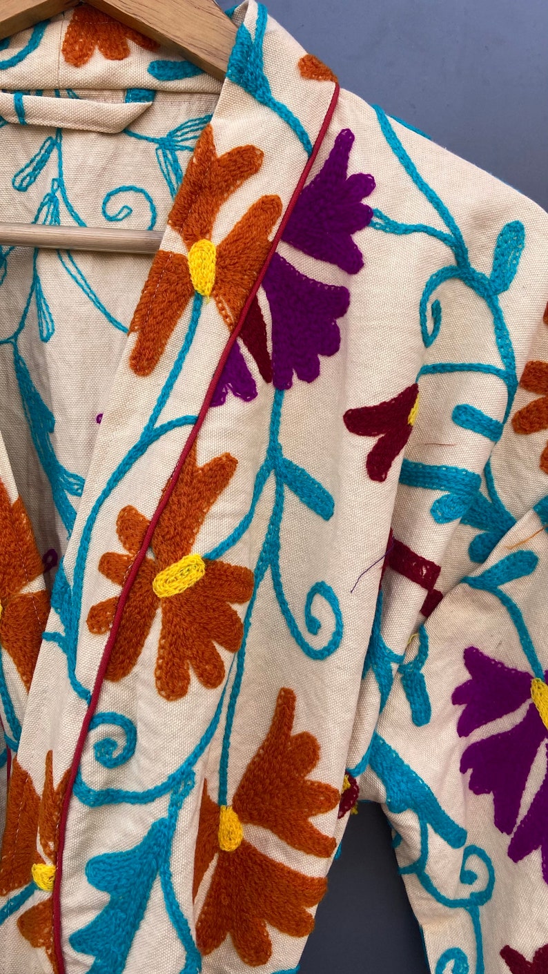 May include: A white cotton robe with a colorful floral embroidery design. The embroidery features turquoise, purple, orange, and yellow thread. The robe has a red trim along the edges.