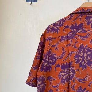 May include: Orange short-sleeved shirt with a purple floral pattern. The shirt is hanging on a wooden hanger. The floral design is a repeating pattern of stylized flowers and leaves.