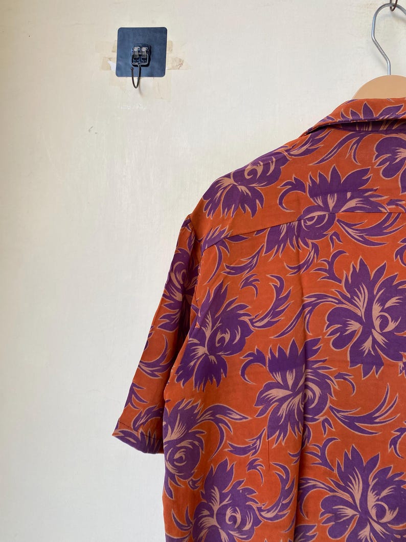 May include: Orange short-sleeved shirt with a purple floral pattern. The shirt is hanging on a wooden hanger. The floral design is a repeating pattern of stylized flowers and leaves.