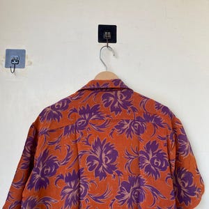 May include: Orange short-sleeved shirt with a purple floral pattern. The shirt is hanging on a wooden hanger. The floral design is a repeating pattern of large, stylized flowers.